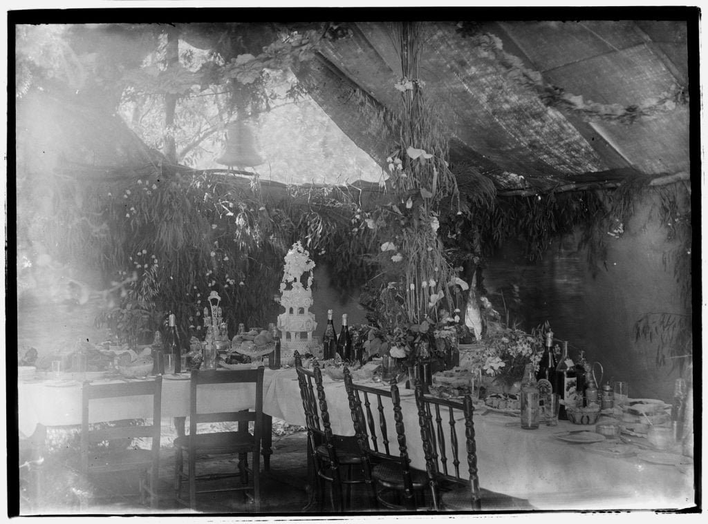 Black and white photograph of a table richly decorated with foliage and laden with bottles, plates, a tiered wedding cake, under a large canopy