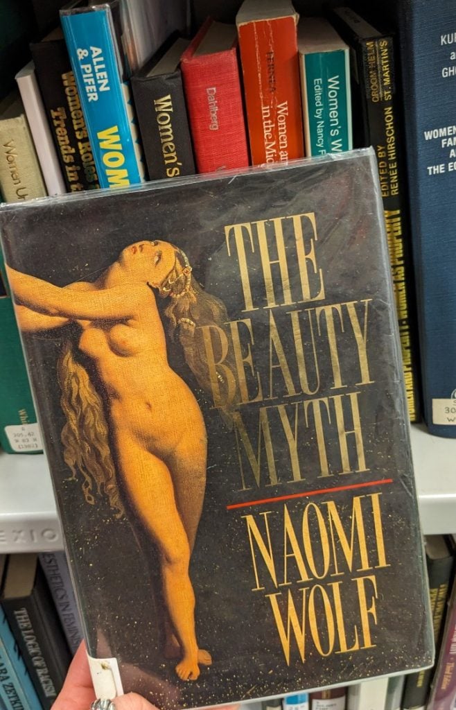 Photo of hand holding a copy of The Beauty Myth by Naomi Wolf in front of two rows of metal bookshelving.