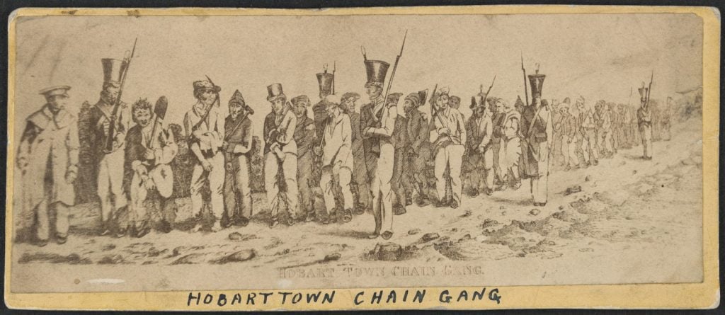 Illustration depicts a line of male convicts, shackled together with leg chains, standing on stony, unmade road. Convicts carry picks, shovels and mallets and wear long pants, jackets and caps; some are barefoot. They are guarded by soldiers wearing military uniforms and carrying rifles.