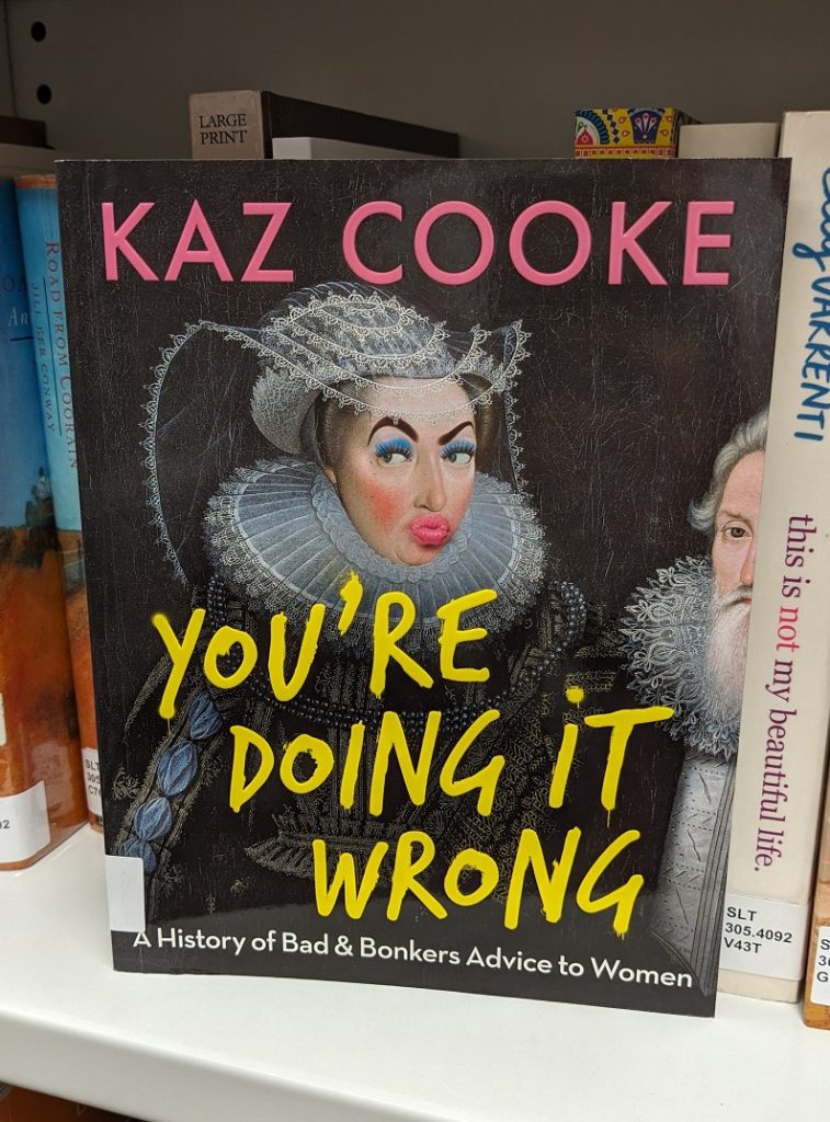 A photograph of the front cover of Kaz Cooke's book 'You're Doing it Wrong', sitting on a metal bookshelf in front of other books. 