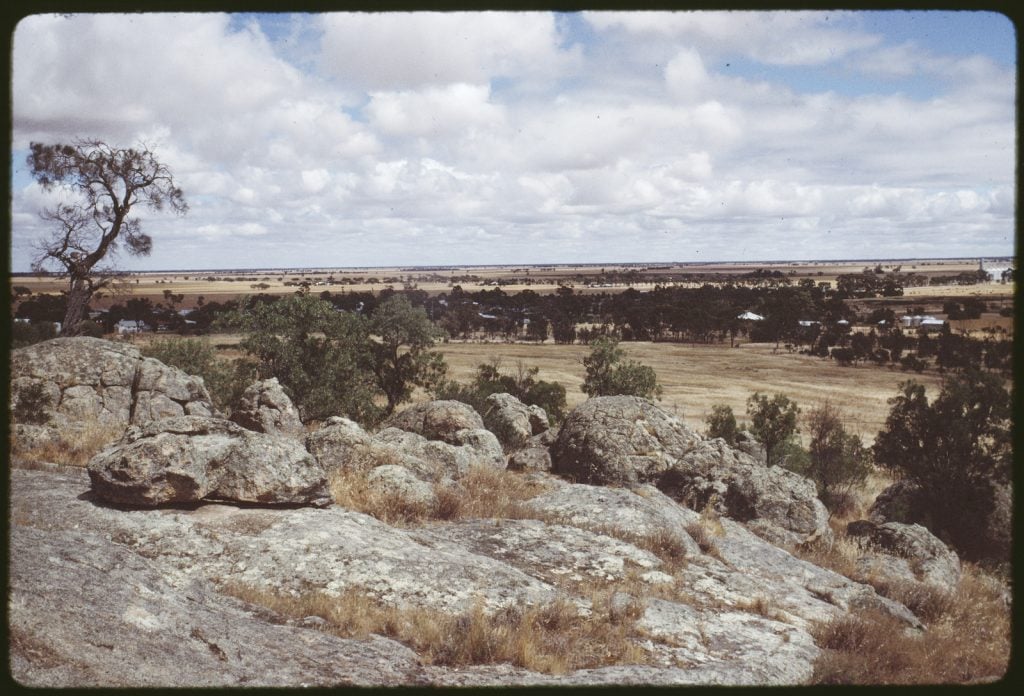 Photo of view from rocks on  Mount Wycheproof. Wide view over plains, showing trees, silos and farm buildings. Background shows wide flat, dry landscape with some trees.    