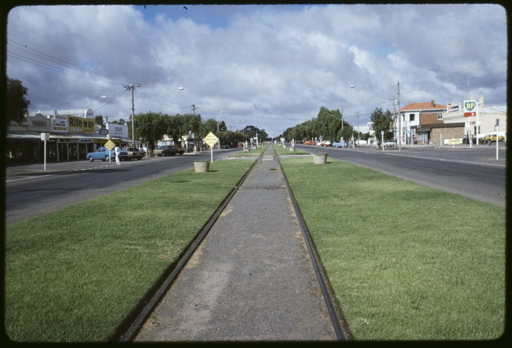 Railway line running down the centre of main street in Wycheproof. Median strip, with roads on both sides. Parked cars in front of shops. Trees in front of houses in distance.  