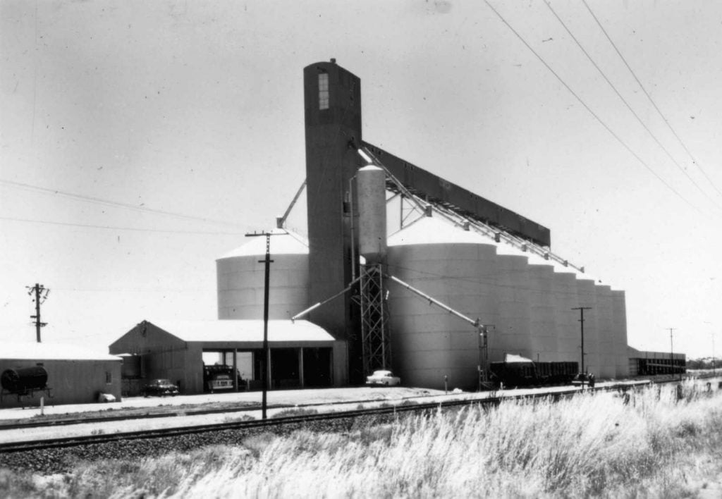 Black and white photo of large grain silos beside two sheds.   Several vehicles parked nearby. Railway line and long dry grass in foreground. 