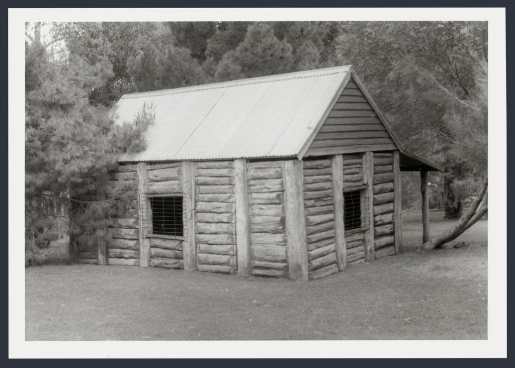 Black and white photo of reconstructed historic wooden house with corrugated iron roof and verandah. Trees behind. Situated in  park at Wycheproof, 1987. 