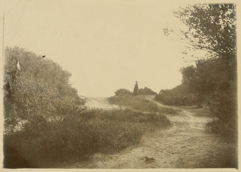black and white photograph of woman dressed in black standing at the end of a sandy track, at the top of the incline.