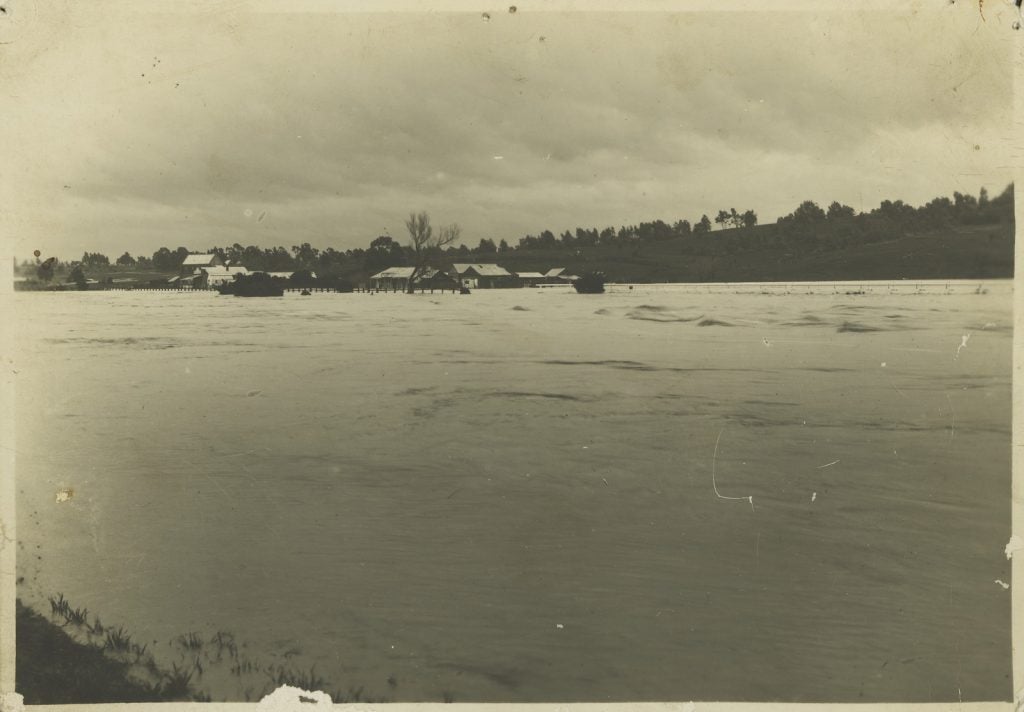 Black and white photograph of view of deep creek in flood, the tops of buildings sitting out o the water in the background