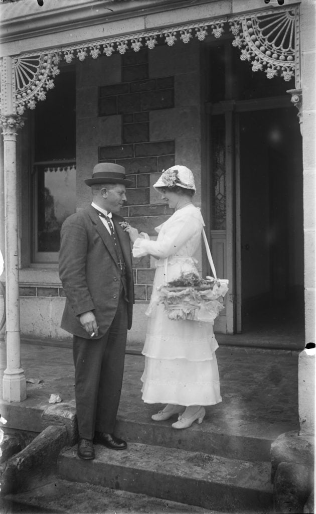 black and white photograph of a man and a woman standing on a front door step, the woman has a basket of corsages over her shoulder and is pinning one onto the man's lapel.