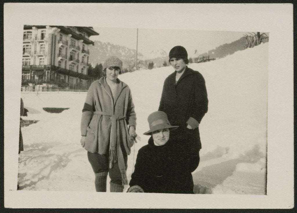 black and white photograph of three women in the snow, a multistory building, possibly a hotel behind.