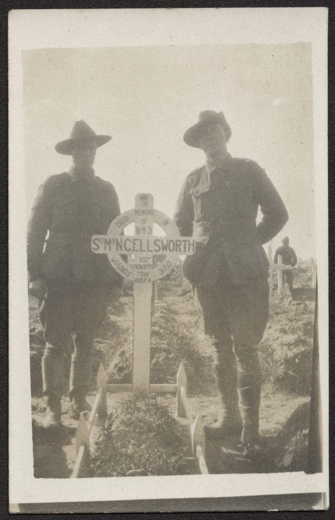 black and white photograph of two soldiers in uniform standing either side of a grave with a cross and white timber edging.