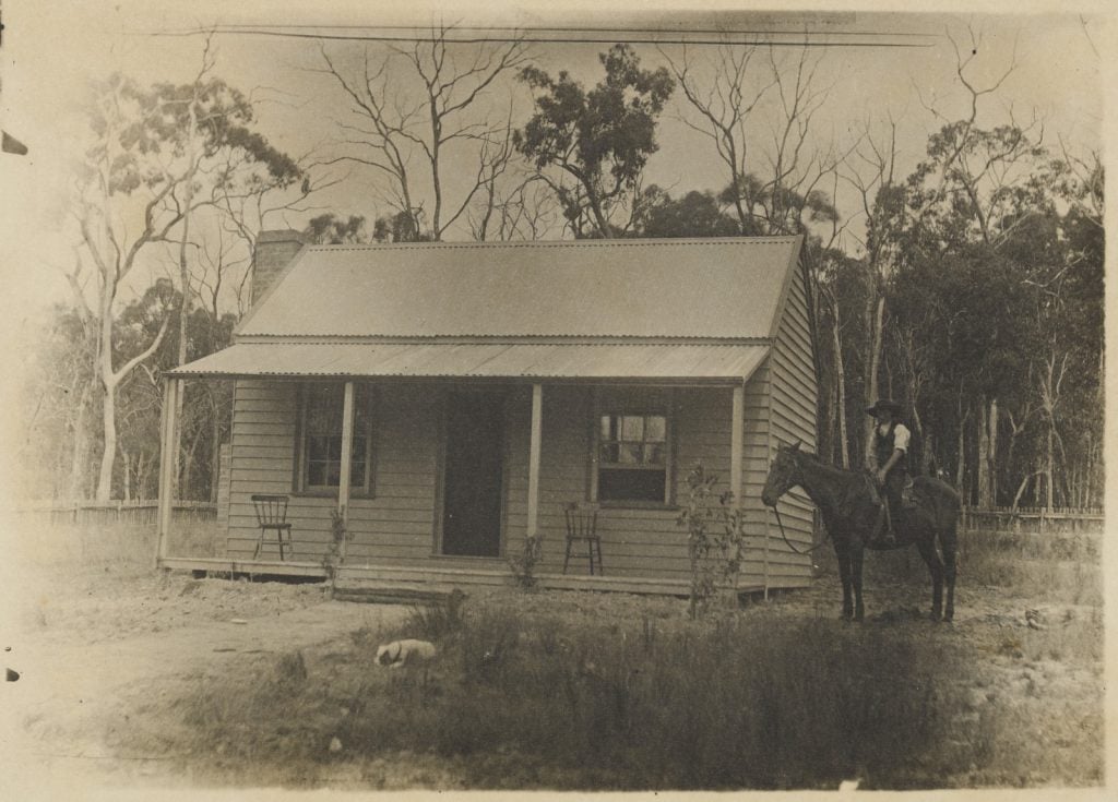 black and white photograph of a cottage, front verandah, with a man on a hosrse beside.