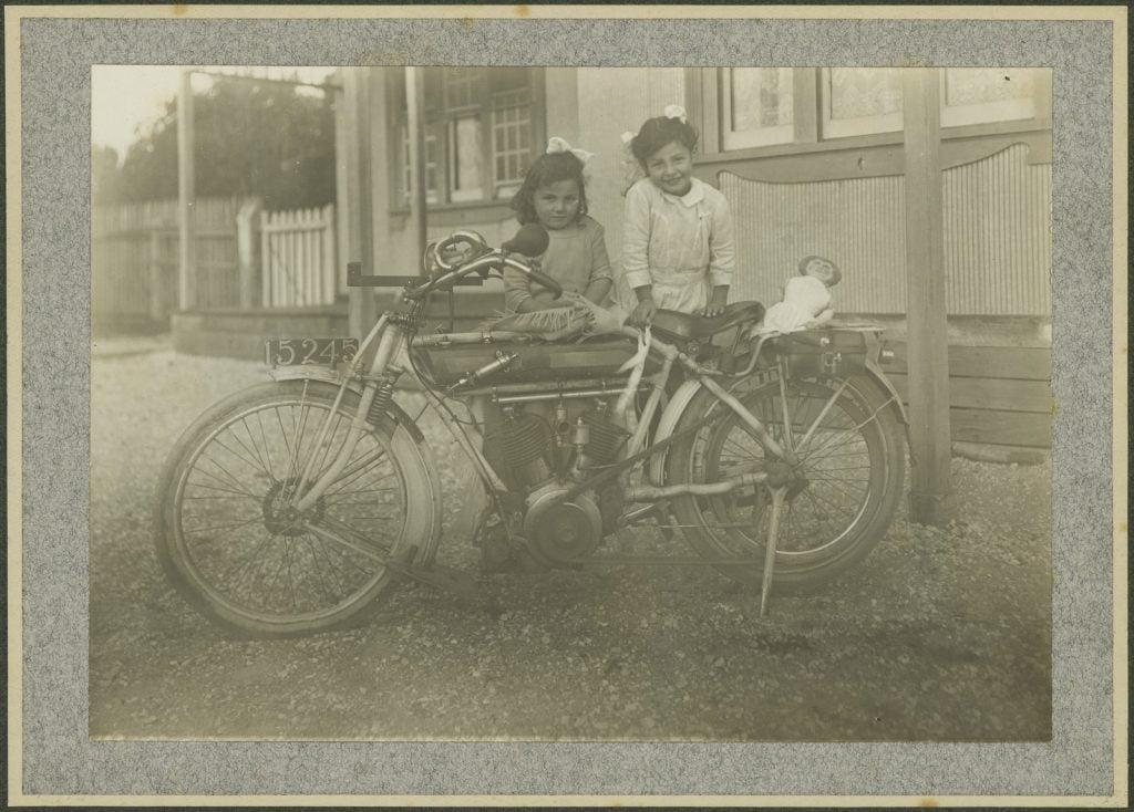 Black and white photograph of two young girls standing beside a motorbike, with a doll as pillion passenger