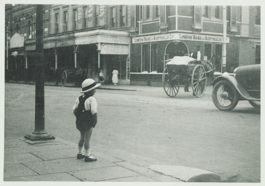 Black and white photograph of a view from behind of young boy in shorts with a straw hat, standing looking across a road with a buggy turning the corner, and the front wheel of a car.