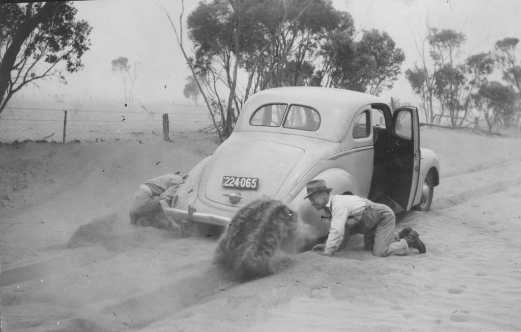 Two men kneel beside a car bogged in sand, trying to dig the sand out from under the wheels. Wycheproof area, December 1944. From The Argus, 29 December 1944, page 7.