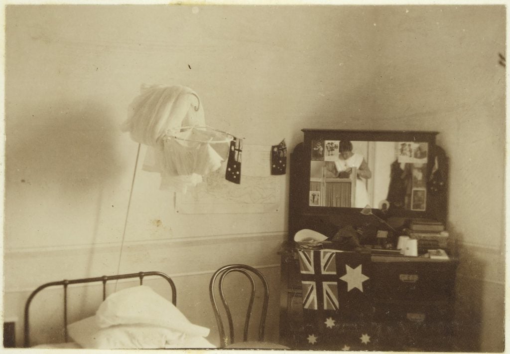 Shows head of bed on left with dressing table with mirror at right, Australian flags suspended from wall and dressing table, nurse seen in mirror's reflection.
