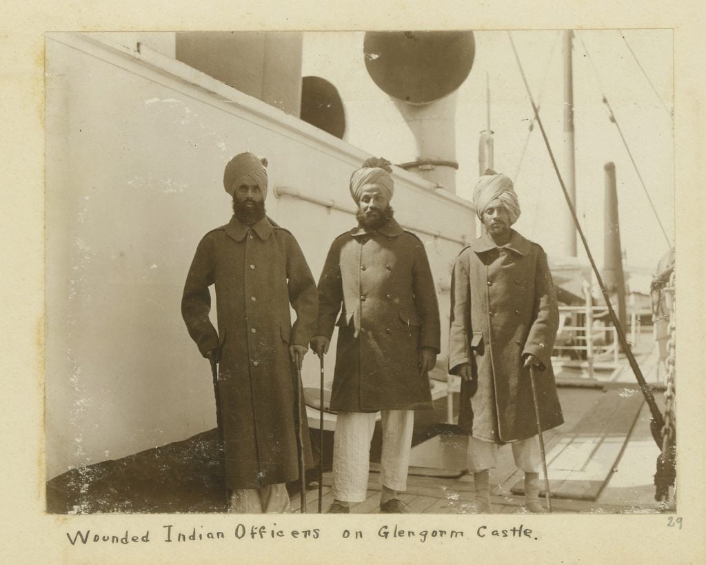 Three Indian soldiers in traditional dress (full-length dark jackets and write pants) and turbans, supported by canes  ship deck.