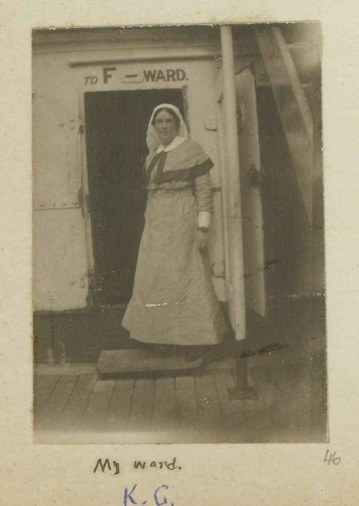 Full-length photograph of female standing to right, wearing ankle length nurses' uniform with cape and veil, Standing at entrance with sign above door reading: To F Ward.