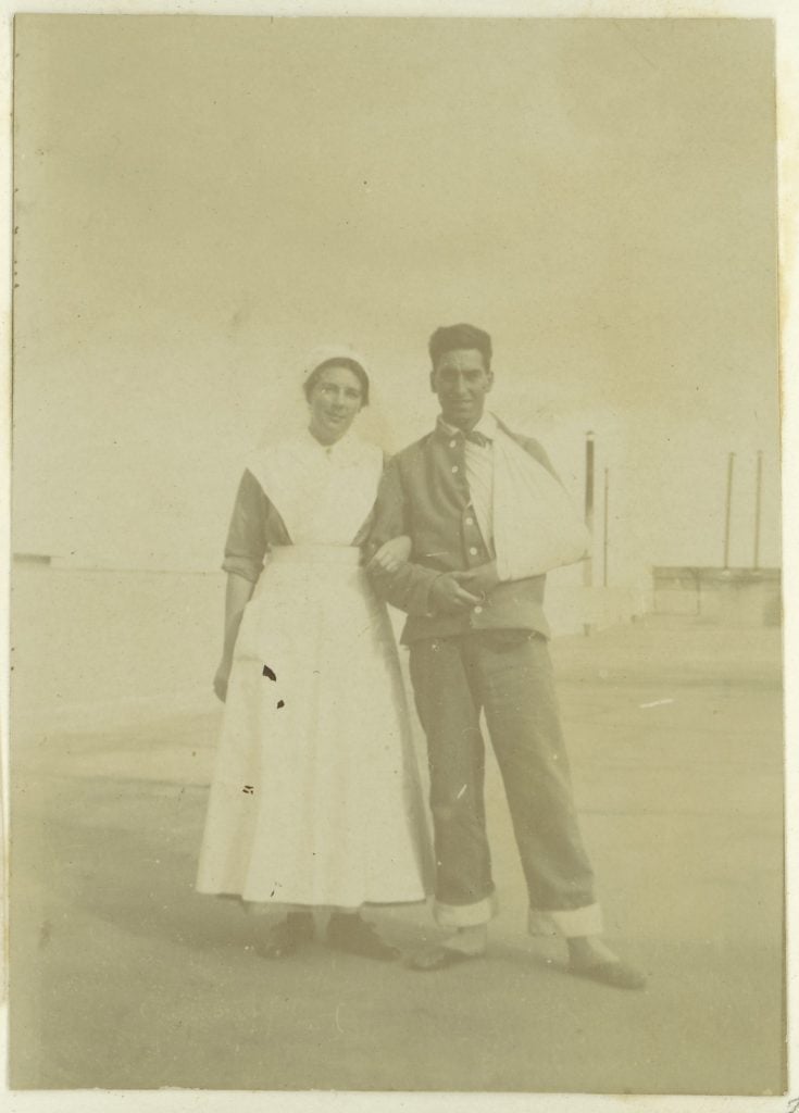 Full-length photograph of nurse wearing ankle length nurses' uniform and apron and veil, standing with arm linked to a soldier with his arm in a sling. The soldier is wearing loose fitting pants with jacket.
