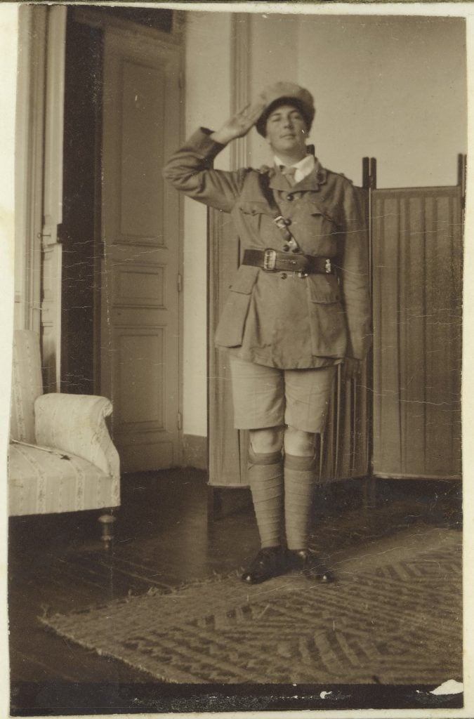 Full-length photograph of female in dressed in a soldier's uniform in a solute pose; long jacket and belt, shorts, full-length socks and leather shoes. In a room with door slightly ajar, lounge chair at right, privacy screen at left, wooden floor covered by a rug. 