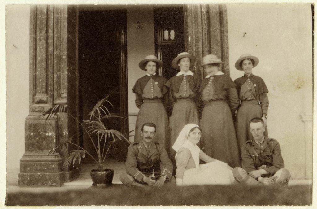 Four nurses standing in formal uniform outside a hotel entrance (full-length dress with shoulder- length cape and hat). A potted plant as well as  two soldiers in uniform sit cross-legged on ground, with nursing Sister in the centre wearing wearing nurses uniform with apron and veil.
