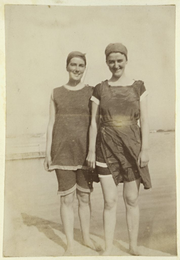 Full-length photograph of two females at the beach, standing at water's edge, both wearing bathing suits composed of a smock over knee length loose pants, with bathing caps.