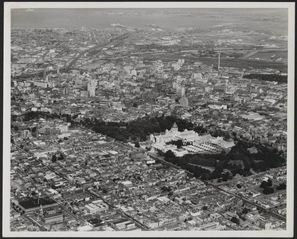 Aerial view of Melbourne looking west with Fitzroy Town Hall in left foreground, Exhibition Buildings and Carlton Gardens with huts middle right, dome of the State Library and uncovered Shot Tower in centre of image, Spencer Street railway lines, Yarra River and Bay in background.