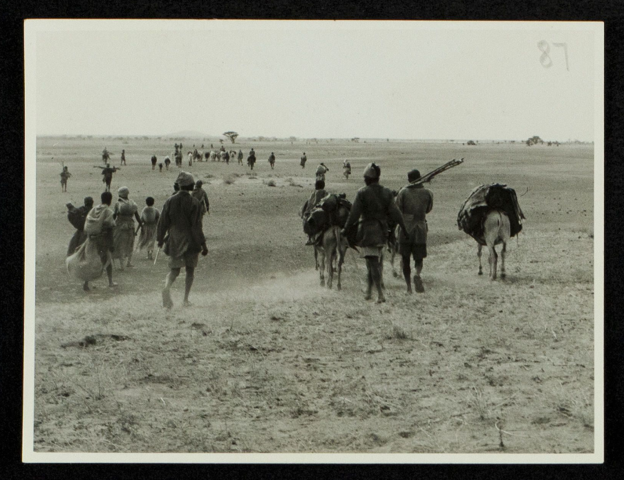 Black and white photograph of people walking across a parched landscape, donkeys carrying belongings.