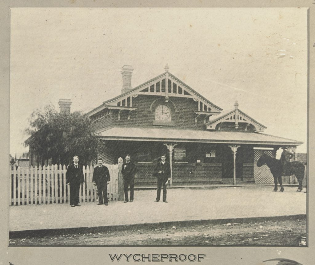 Black and white photo of Wycheproof post office, a brick building with verandah and large clock on front. Picket fence at left. Four men stand in front of the post office and one man sits on a horse.  Date is about 1897 to 1899. 