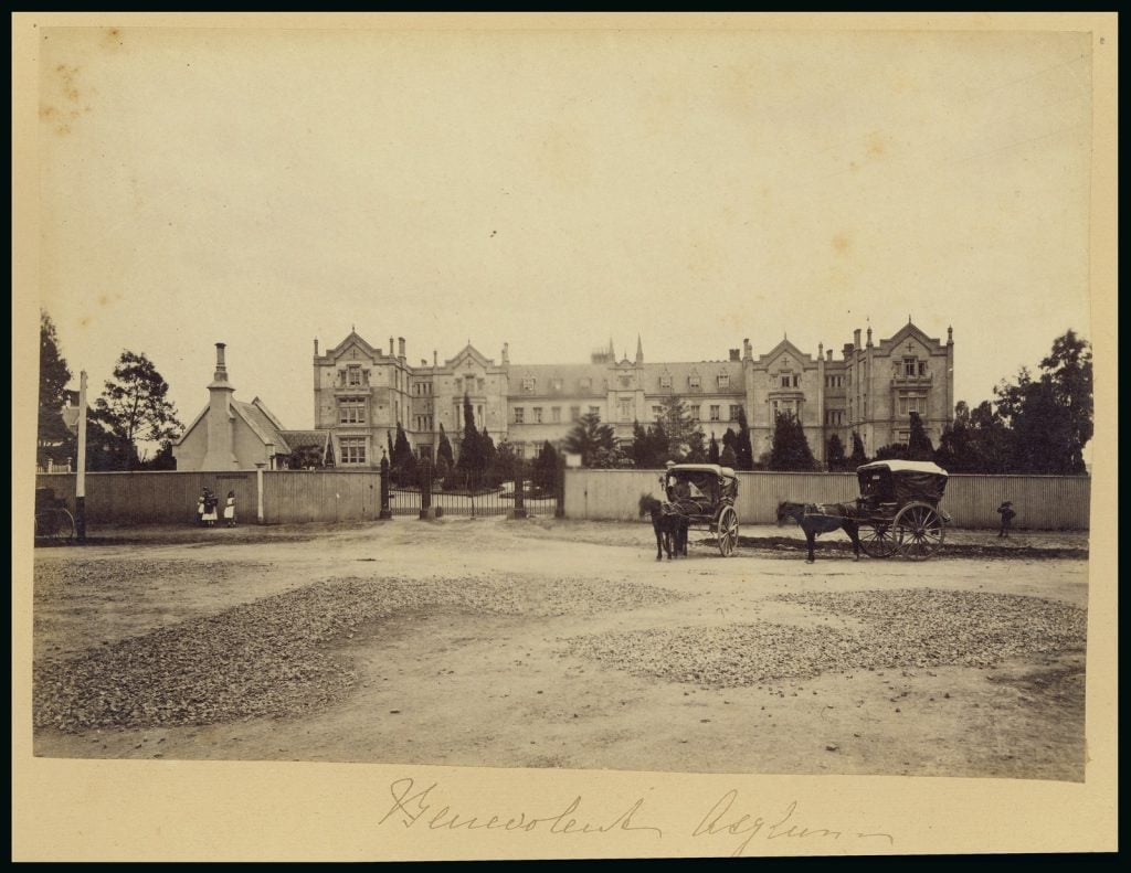 Black and white photograph of large Benevolent Asylum building with horse drawn carts in front and a group of children playing outside the front gates.