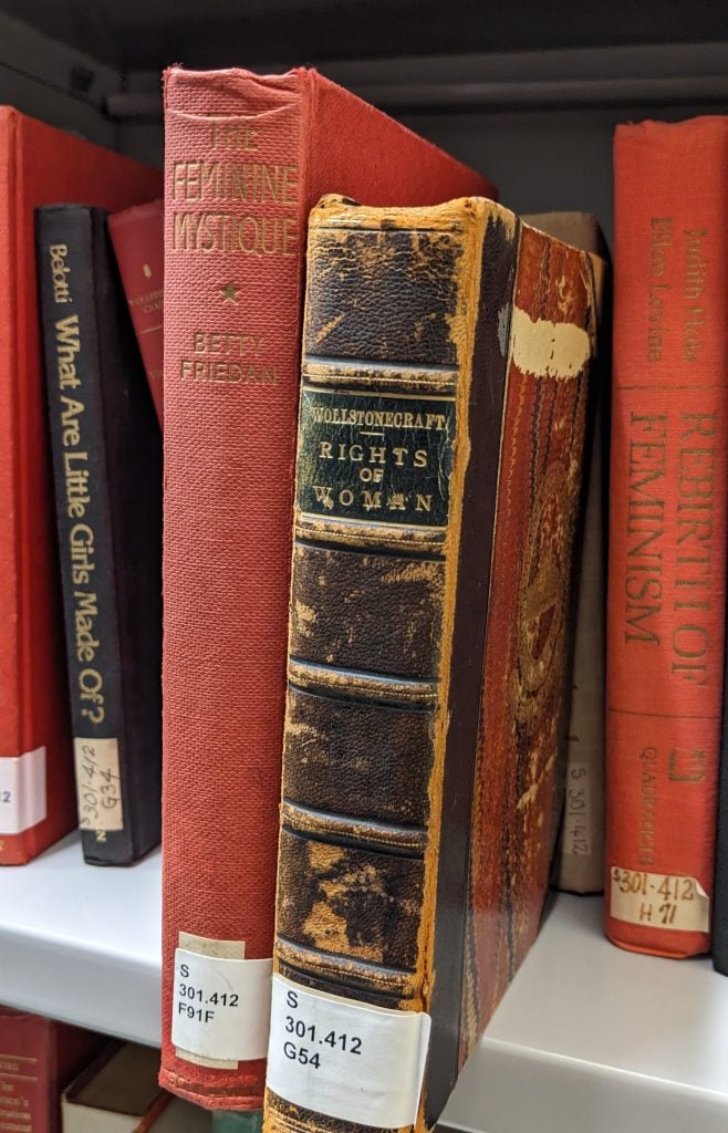 Photograph of Library Stack shelving with two books pulled forward towards the camera so the titles on the spines can be read. One is The Feminie Mystique by Friedan, and the other is Wollstonecraft's Rights of Woman. 