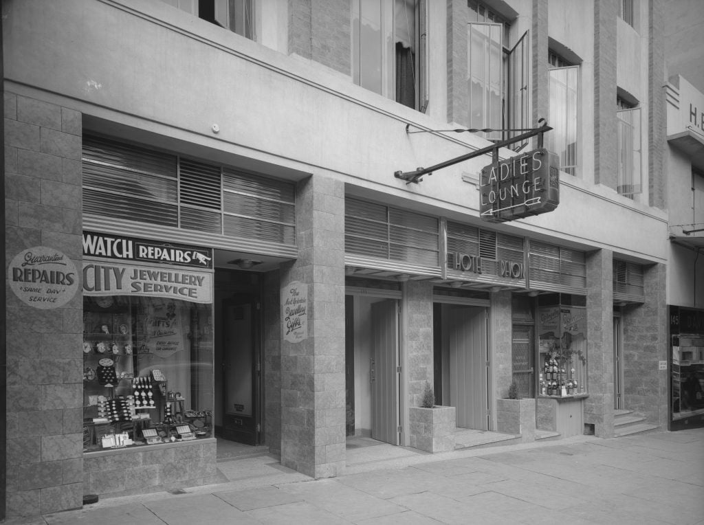 Black and white photo of exterior of Hotel Devon from street level. there is a Neon sign advertising the ladies lounge out the front