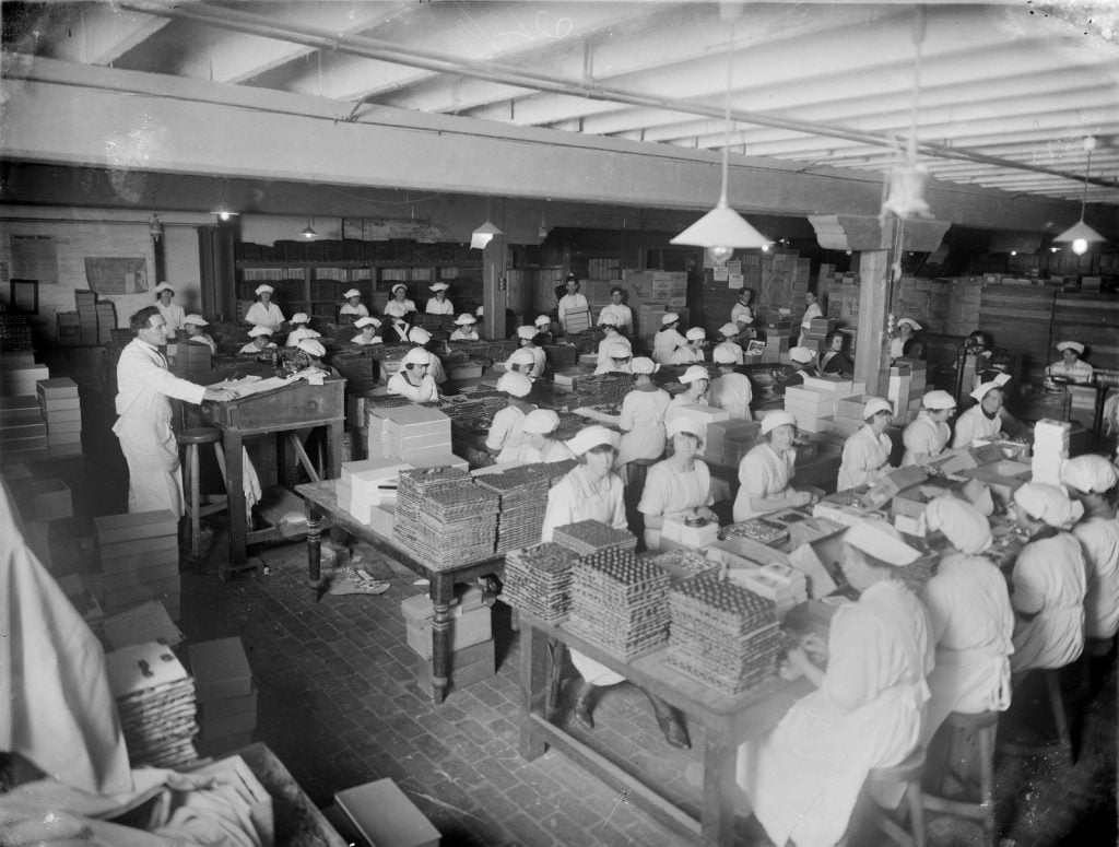 black and white photograph of women dressed in white dresses and aprons sitting at tables packing chocolates into boxes