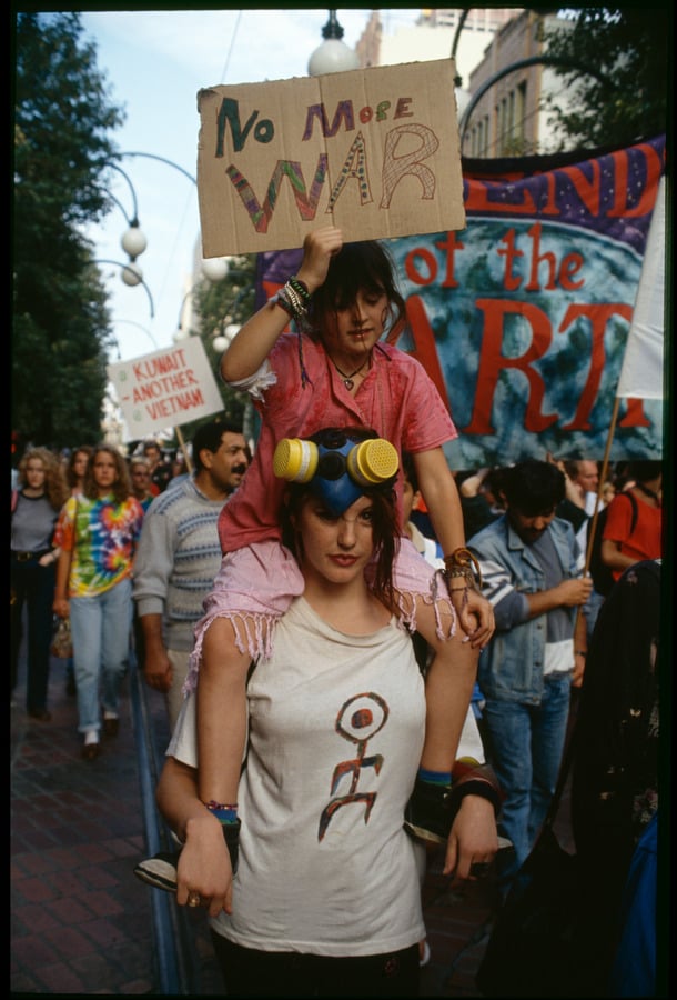 Woman walking in protest march with child sitting on her shoulders, holding a sign saying 'no more war'