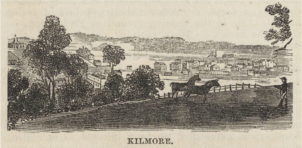 View of the township of Kilmore from a hilltop. There are horses and a man standing in a paddock in the foreground. Gum trees can be seen to the left of the paddock.