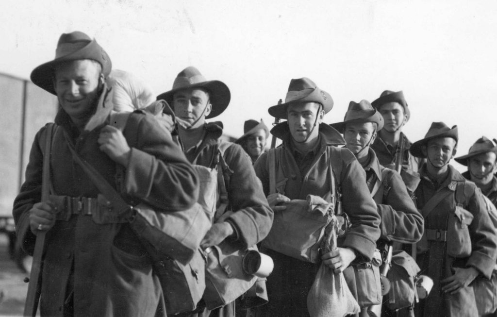 Shows a group of soldiers, wearing slouch hats and carrying their kit bags