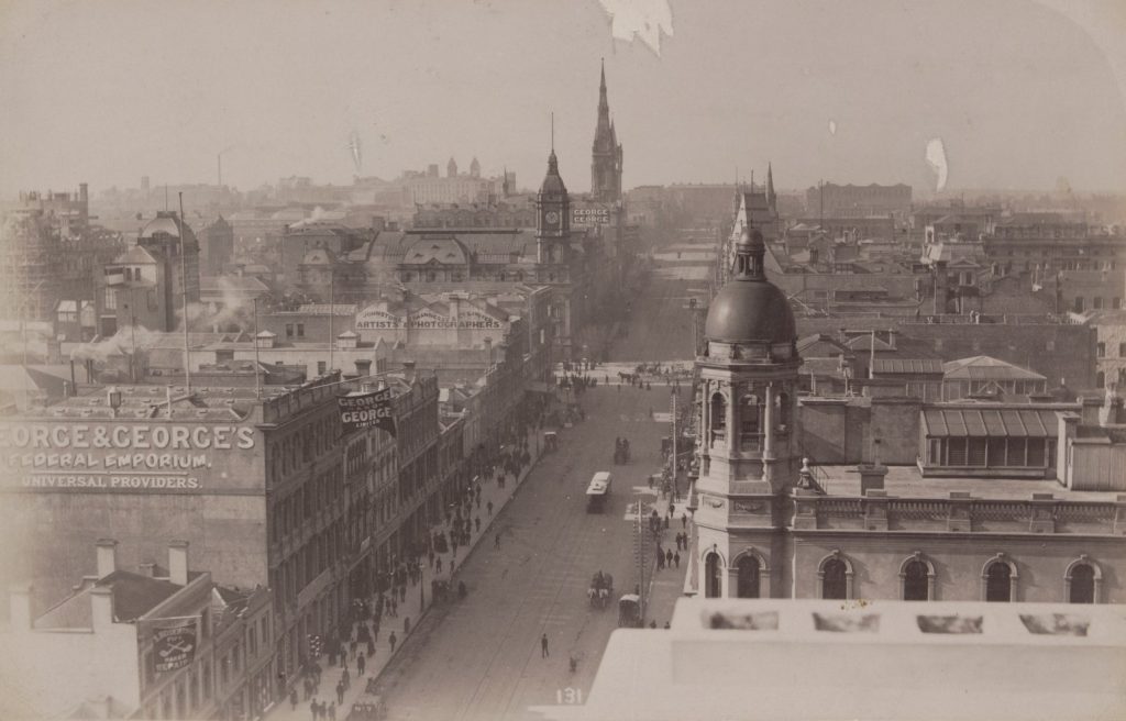 Photograph of Collins Street, Melbourne, elevated view looking east, ca.1890. Shows buildings including Melbourne Town Hall, George & George's Federal Emporium, Johnstone & O'Shannessy Artists & Photographers and the Scots Church spire, as well as horse drawn vehicles, trams and pedestrians. Inscribed in white ink l.c.: 131.