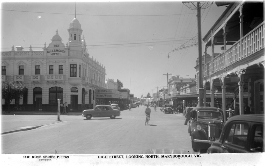 Black and white view of High Street, Maryborough. There are old cars parked on the side of the road, pedestrians walking along he shopping strip and a person on a bicycle riding along the street. The Bull and Mouth Hotel is on the street corner to the left of frame.