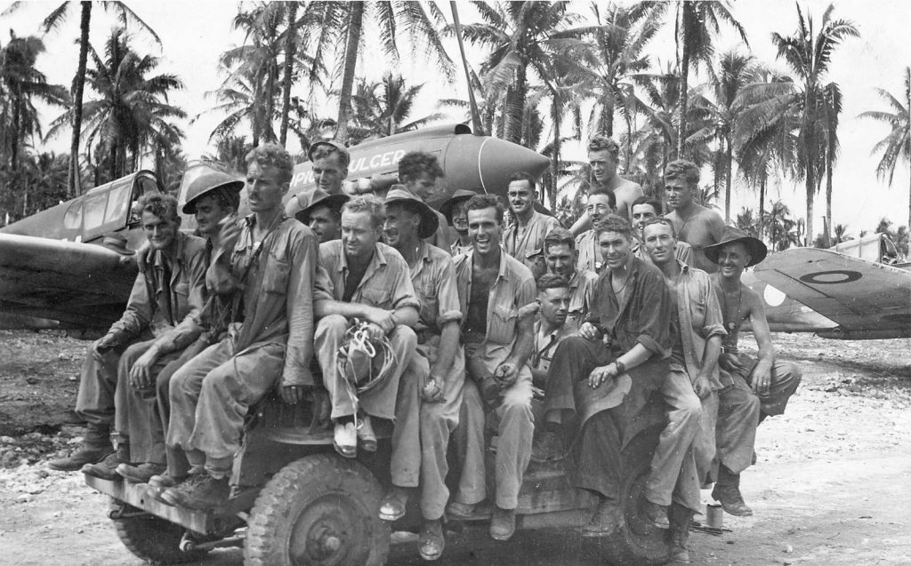 Shows 20 service men in working clothes, seated on a tank. two aircraft are visible in the background.