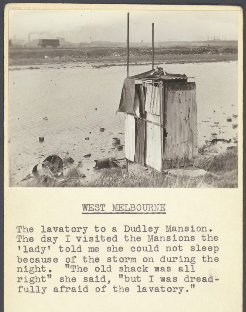 Black and white photo of a
poorly constructed lavatory building on the edge of a swamp. Construction material is mainly corrugated iron. Rail freight car and industrial landscape in the background. Typewritten caption underneath reads 'The lavatory to a Dudley Mansion. The day I visited the Mansions the 'lady' told me she could not sleep because of the storm on during the night. "The old shack was alright" she said, "but I was dreadfully afraid of the lavatory."