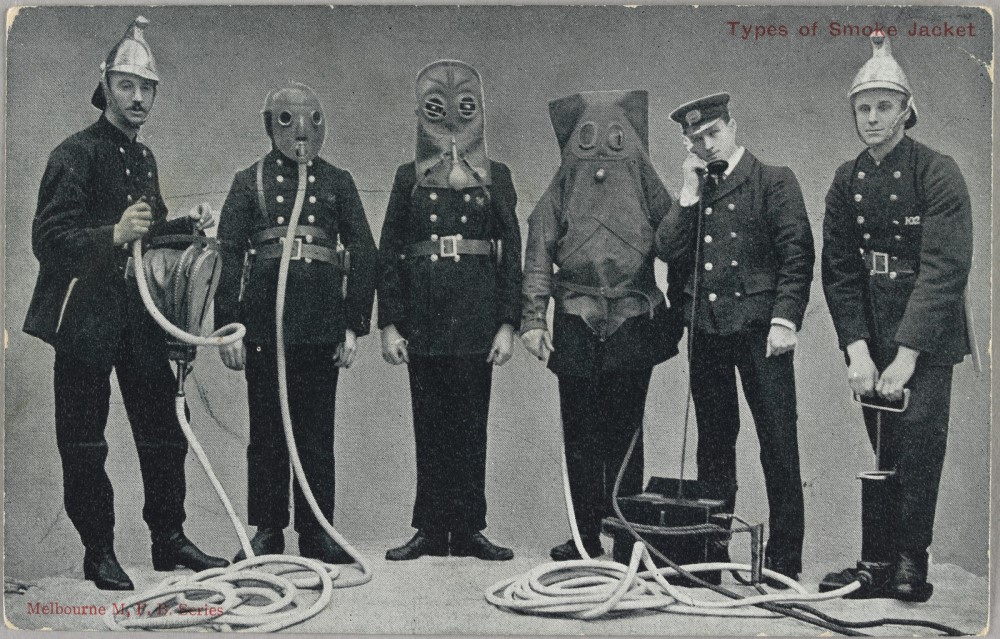 Line of six uniformed firemen, three wearing different types of smoke jacket masks. Two firemen wearing helmets show different pumps with hoses connected to two masks. One fireman listens to telephone hand piece connected to a mask.