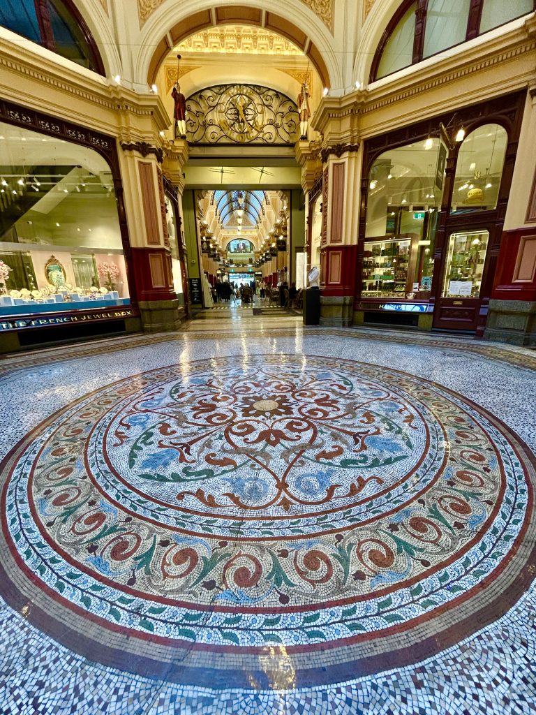 Photo of a section of the Block Arcade with mandala circular mosaic flooring prominently featured. Arcade shopfronts are visible in the background, and arched walkway.