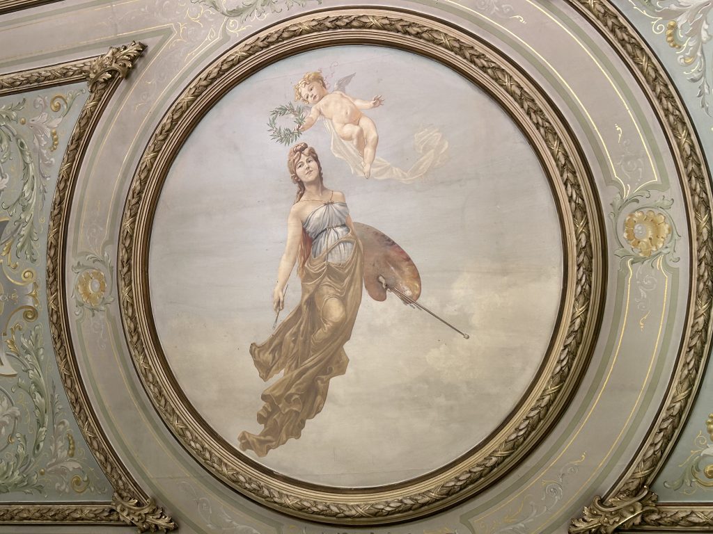 Circular ceiling mural showing a angel in loose flowing garments and a small childlike winged cherub, surround by decorative gold-edging and floral decorative borders.
