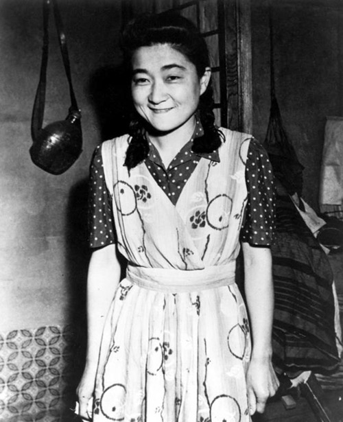 Iva Toguri stands smiling next to her radio microphone. She wears a brightly patterned dress.