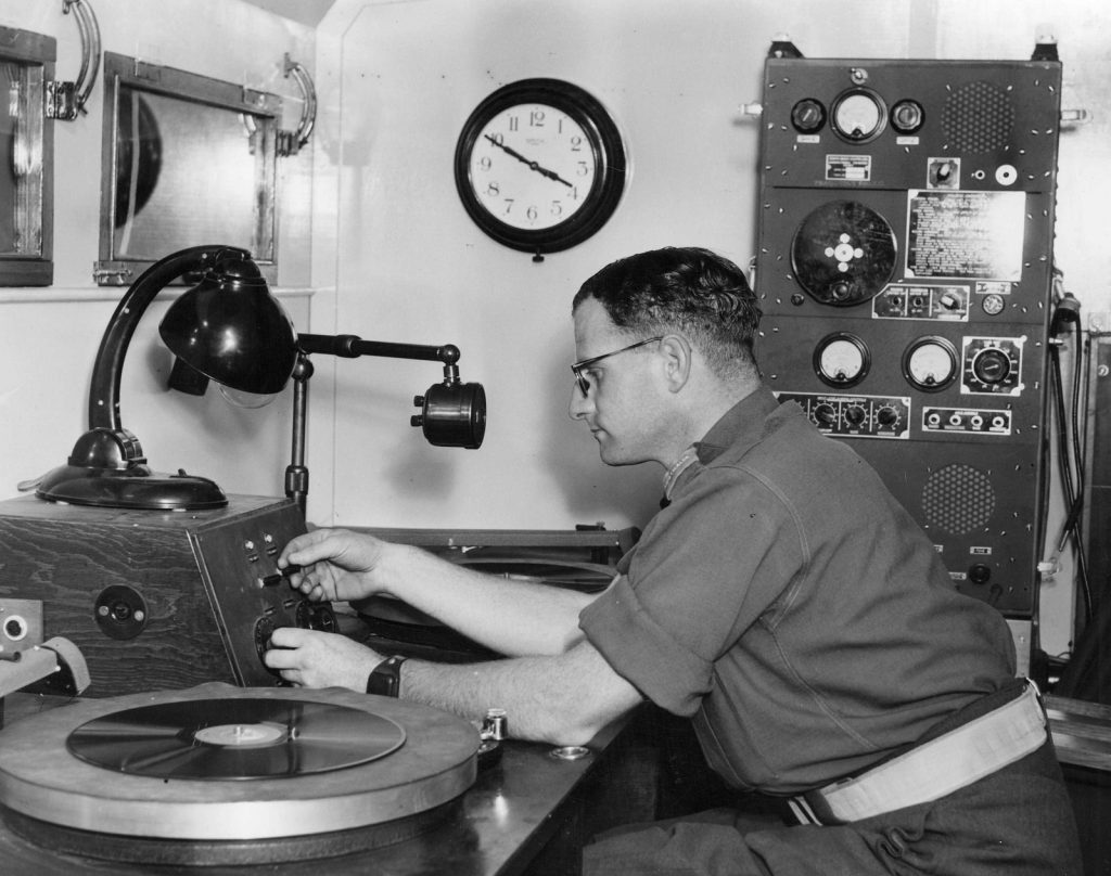 Sergeant in Australian Army sits at radio microphone and turntable. Radio machinery at side. Clock on wall.