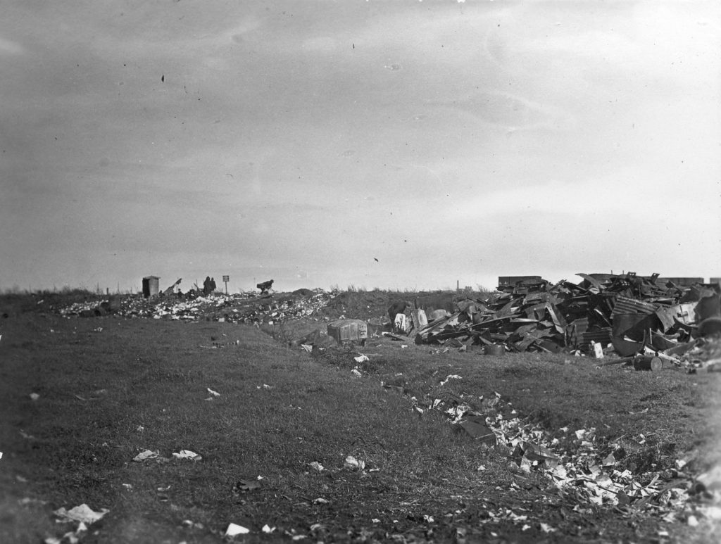 Black and white photo of open area strewn with rubbish. Three figures are visible on the horizon.