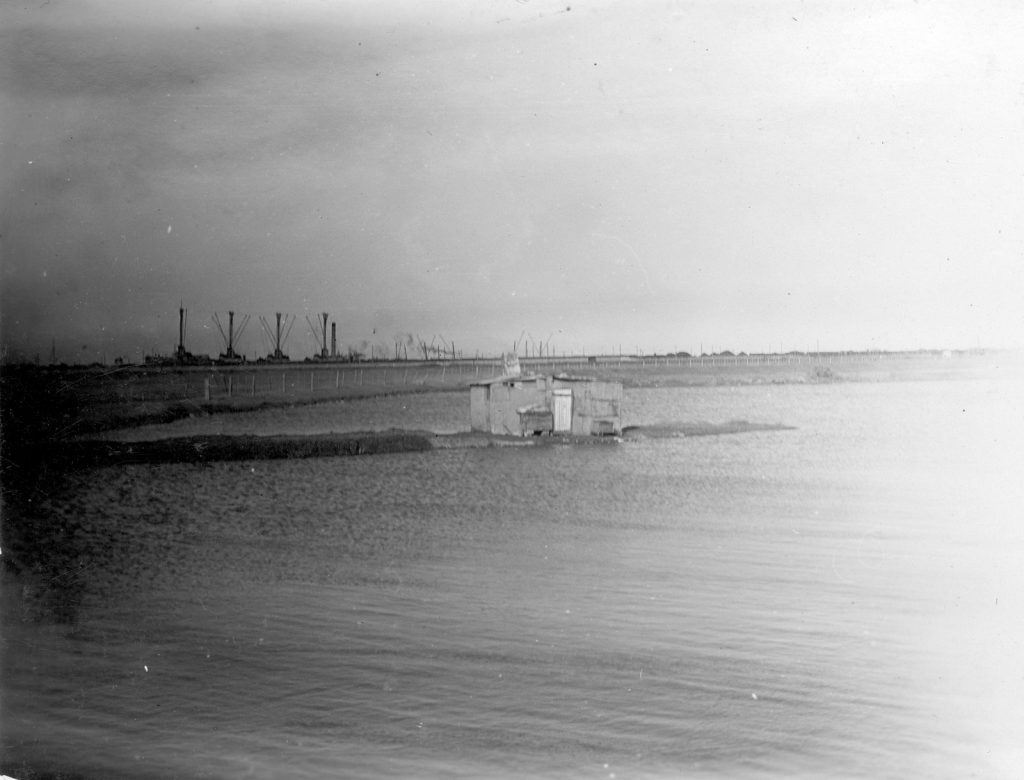 View across a stretch of water to a shanty built on a spit. More water and industrial landscape in the background.