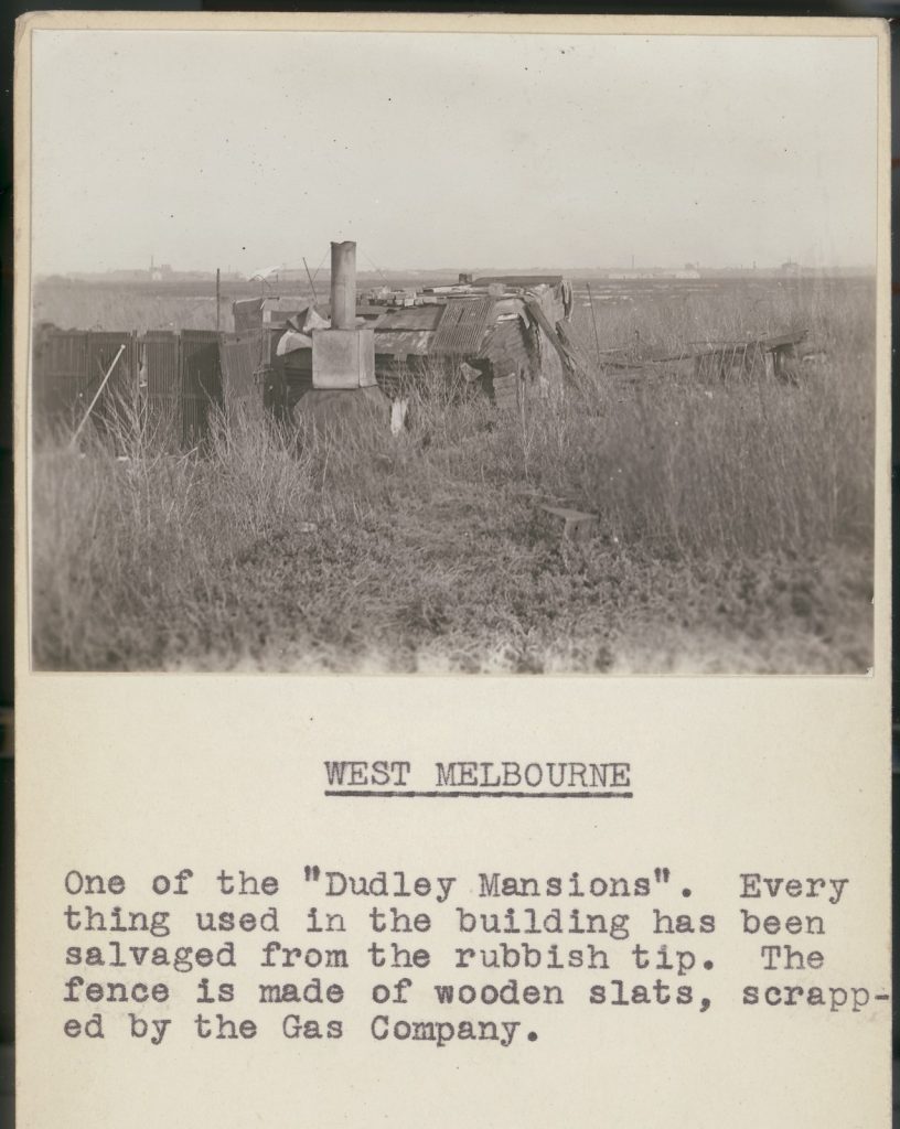 Picture of a dwelling made out of rubbish. Typewritten caption underneath reads 'One of the "Dudley Mansions". Everything used in the building has been salvaged from the rubbish tip. The fence is made of wooden slats, scrapped by the Gas Company.