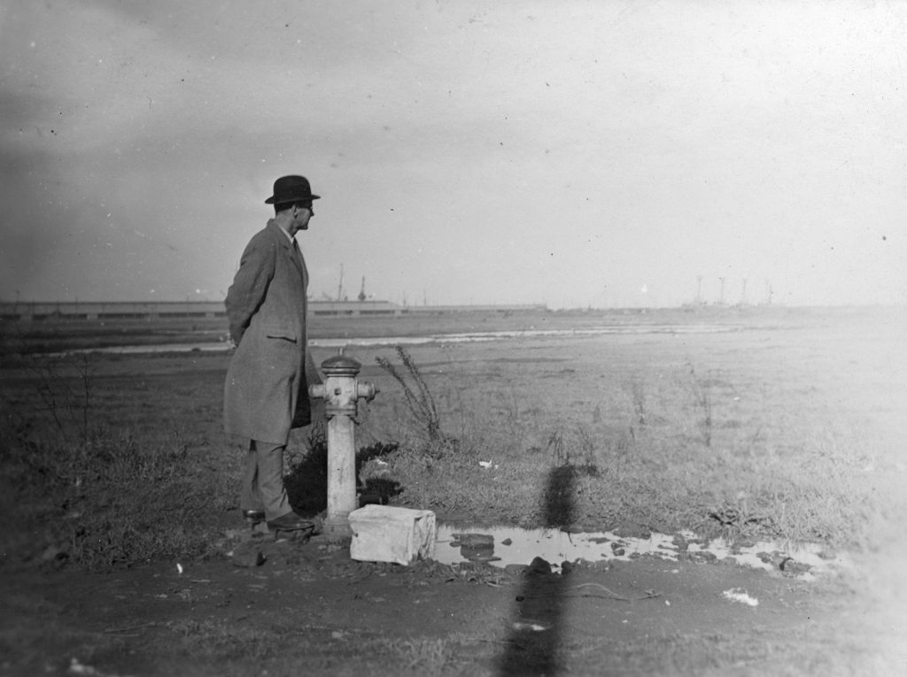 Black and white photo of a man
dressed in a bowler hat and overcoat standing next to a water hydrant in a large open area. Water is visible on the ground. There is an industrial landscape in the background.