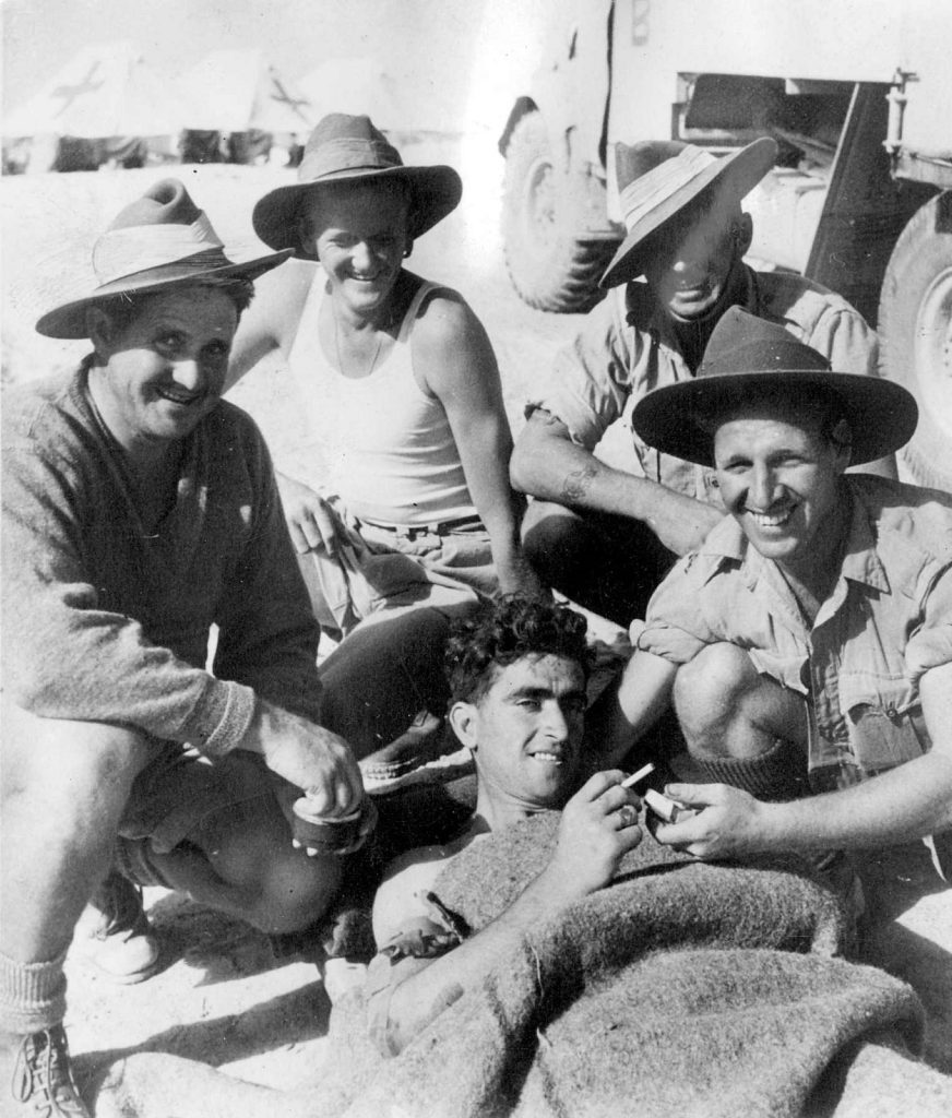 Four soldiers crouched on the ground wearing slouch hats. One in the foreground, laying down and holding a cigarette.