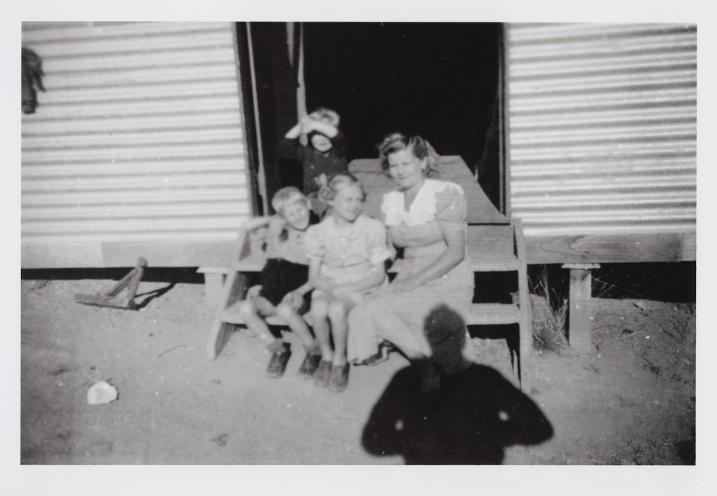 Black and white slightly blurry photograph of woman and three children sitting on the steps of a hut made of corrugated metal, in the shade of a male figure, presumably taking the photograph.