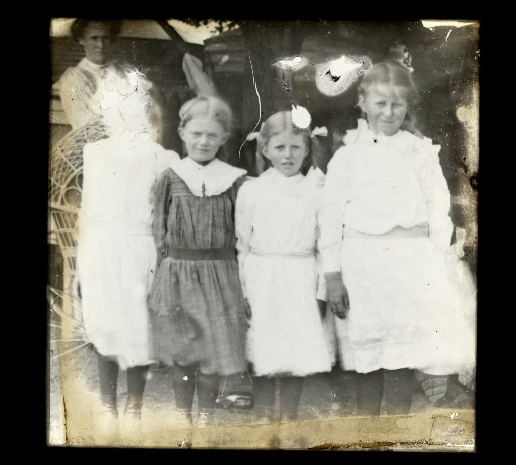 Photograph of four young girls wearing knee length dresses. Face of girl on left obliterated by emulsion damage.